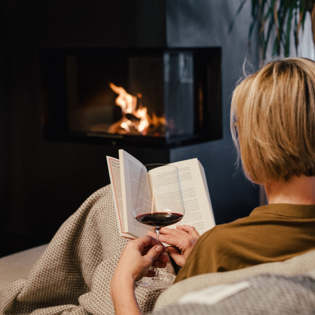 Frau liest am Kamin unter einer Decke, in der Hand ein Glas Rotwein