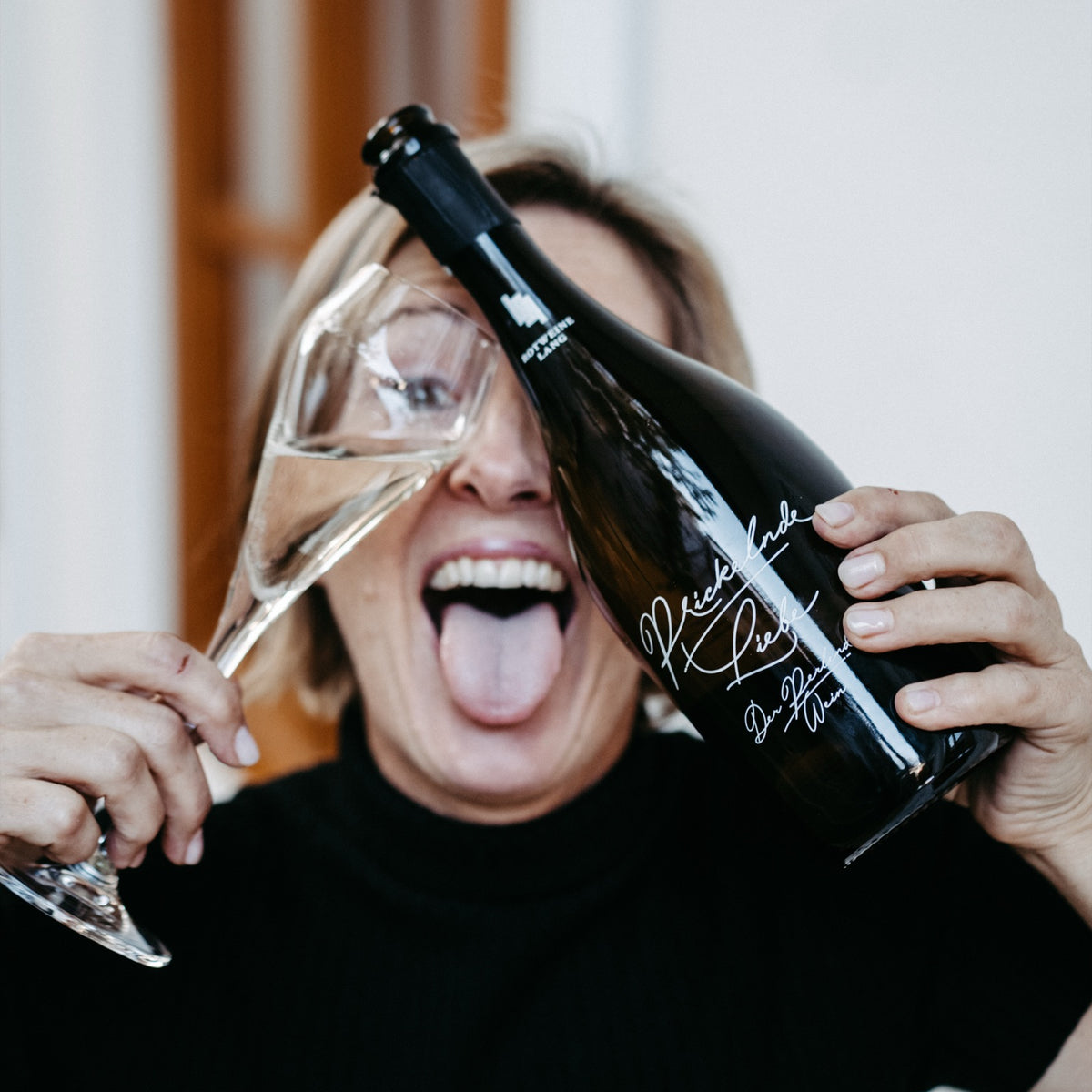 Andrea Lang mit einer Flasche Prickelnde Liebe und einem Glas Frizzante, verschmitzt mit herausgestreckter Zunge